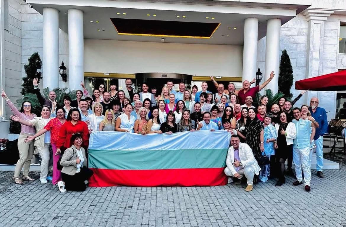 Голяма група партньори от екипа с българското знаме пред хотел, в слънчев ден